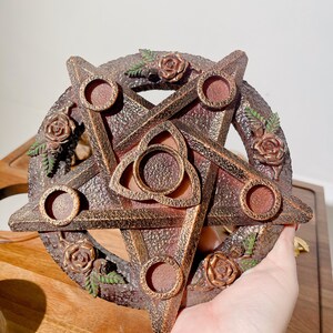 May include: A circular pentagram candle holder with a textured bronze finish. The design features five small circular candle cups at each point, a central triquetra symbol, and decorative sculpted roses with green leaves around the perimeter.