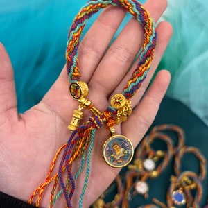 May include: A colorful braided bracelet with gold accents and a round charm featuring a painted image of a person. The bracelet has tassels in red, blue, and green.