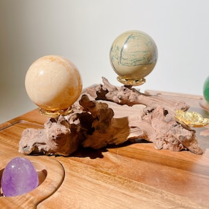 May include: Two polished stone spheres, one light orange and one sage green with dark veining, rest on gold-colored metal flower-shaped stands attached to a piece of natural driftwood. A smooth purple stone sits in a circular cutout on a wooden base.