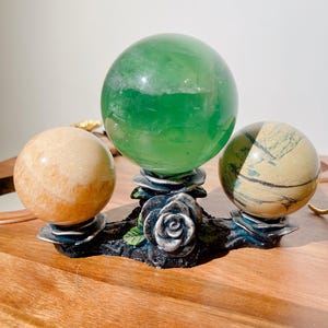 May include: A decorative stand featuring three silver-colored metal roses with green leaves, holding three polished stone spheres. The central sphere is translucent green, while the two side spheres are opaque tan and beige with dark veining.