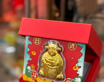 Peut inclure: Une figurine dorée d'une divinité chinoise tenant un lingot d'or, dans une boîte rouge avec un motif or et rouge. La boîte porte les caractères chinois "日进" (ri jin) qui signifie "progrès quotidien" et "金玉满堂" (jin yu man tang) qui signifie "une maison pleine d'or et de jade".