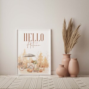 Peut inclure: Une impression encadr&eacute;e blanche avec le texte "HELLO Autumn" en lettres brunes. L'impression pr&eacute;sente une illustration aquarelle d'une sc&egrave;ne d'automne avec un v&eacute;lo, des citrouilles et un petit stand avec un parapluie blanc.