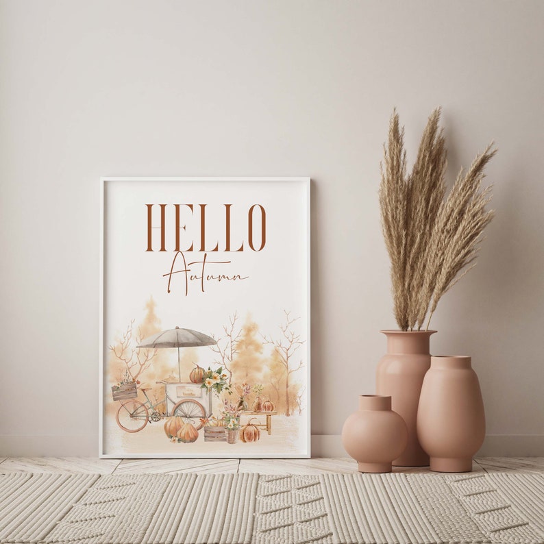 Peut inclure: Une impression encadr&eacute;e blanche avec le texte "HELLO Autumn" en lettres brunes. L'impression pr&eacute;sente une illustration aquarelle d'une sc&egrave;ne d'automne avec un v&eacute;lo, des citrouilles et un petit stand avec un parapluie blanc.