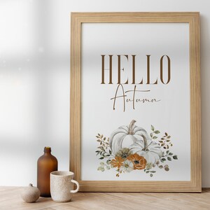 May include: A framed print with the text "HELLO Autumn" in brown lettering. The print features a watercolour illustration of two white pumpkins with orange and white flowers and green leaves.