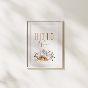 May include: A white framed print with the text "HELLO Autumn" in brown lettering. The print features a watercolour illustration of two white pumpkins with orange and white flowers and green leaves.