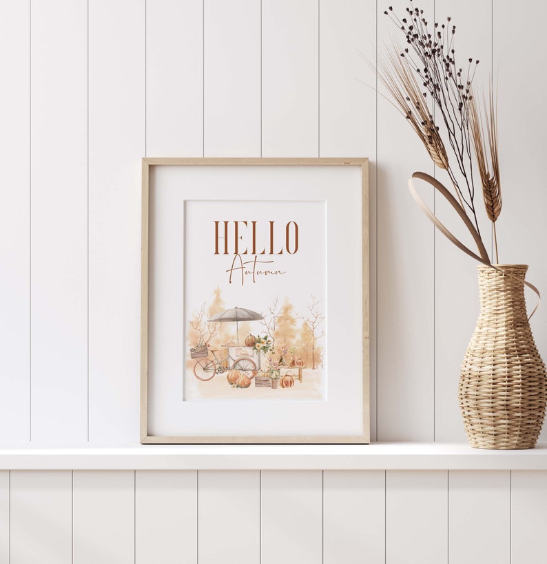 Peut inclure: Une impression encadr&eacute;e avec le texte "HELLO Autumn" en lettres brunes. L'impression pr&eacute;sente une illustration aquarelle d'un v&eacute;lo avec une verri&egrave;re, des citrouilles et des feuillages d'automne.