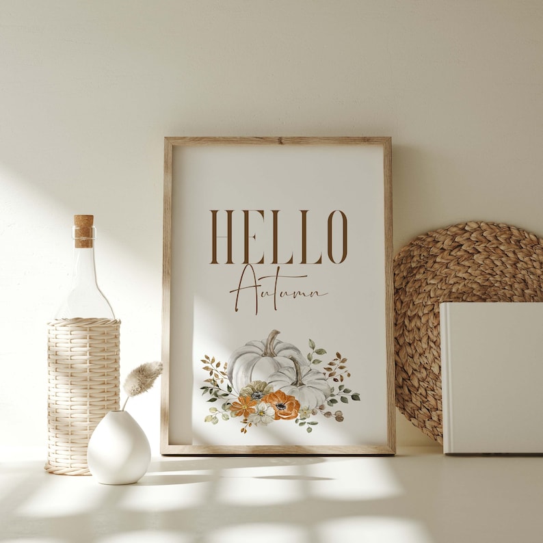 May include: A framed print with the text "HELLO Autumn" in brown lettering. A white pumpkin with orange and white flowers is depicted below the text.