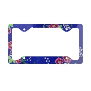 May include: A blue license plate frame with a floral design featuring pink and white flowers. The frame has two holes on each side for attaching to a license plate.