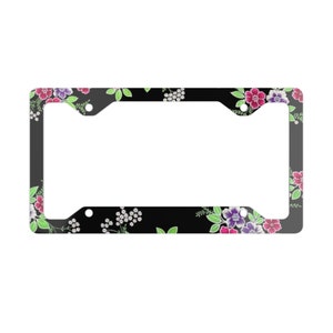 May include: Black license plate frame with a floral design. The frame features a pattern of pink, purple, and white flowers with green leaves. The flowers are set against a black background.