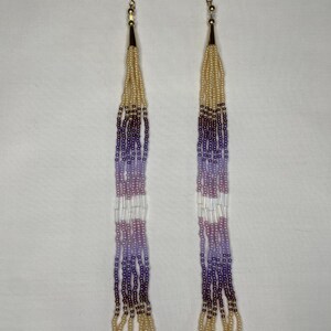 May include: A pair of long beaded earrings with gold-colored hooks and accents. The earrings feature a gradient of colors, including cream, purple, and white beads. The bottom of the earrings have multiple strands of cream-colored beads.