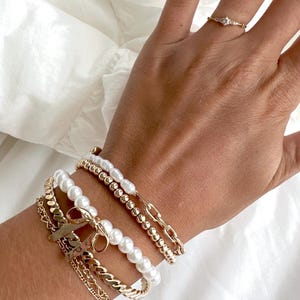 Puede incluir: Un conjunto de cuatro pulseras que incluyen una pulsera de cadena dorada, una pulsera de cadena dorada con un pequeño dije de lazo y dos pulseras de perlas.