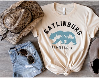 May include: A beige t-shirt with a black graphic of a bear silhouette with mountains and trees inside. The text "Gatlinburg Tennessee" is printed below the bear.