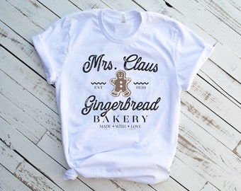 May include: White t-shirt with a black and brown graphic design that says "Mrs. Claus Gingerbread Bakery Est. 1930 Made with Love". The graphic includes a gingerbread man.