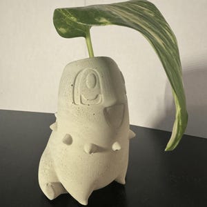 Könnte beinhalten: Ein weißer Betontopf in Form eines Cartoon-Monsters mit Stacheln und einem Blatt, das aus der Oberseite herausragt.