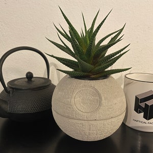 Peut inclure: Un pot en céramique blanche en forme d'Étoile Noire de Star Wars, avec une plante succulente verte qui pousse à l'intérieur. Une théière en fonte noire et une tasse blanche avec le texte "Haptical Factory" sont également présents sur l'image.