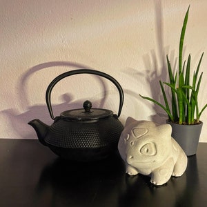 Könnte beinhalten: Eine schwarze gusseiserne Teekanne mit einem runden Körper und einem Ausguss an der Seite. Eine weiße Keramikfigur eines Comic-Charakters mit einem großen Kopf und einem breiten Lächeln befindet sich im Vordergrund. Eine Topfpflanze in Grün befindet sich im Hintergrund.