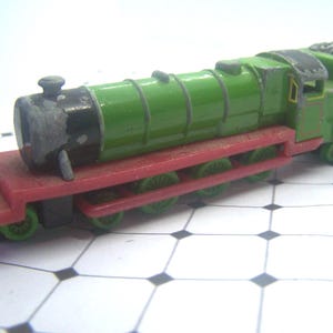 May include: A toy train featuring a green engine and coal car, mounted on a red base with green wheels. The train has black accents and is set against a white and black checkered background. A classic toy.