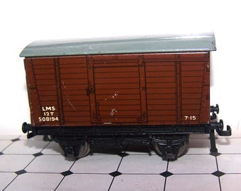 May include: A brown model railway boxcar with a grey roof and black undercarriage. The side features white lettering that reads LMS, 12 T, 508194, and 7-15. The model includes metal wheels and buffers, showcasing a detailed planked wood texture.