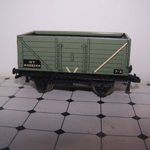 May include: A miniature olive green model train freight car with black wheels and undercarriage. The car has a rectangular shape with vertical wooden planks and a white diagonal support beam. The text "12T M608344" and "7-3" are visible.