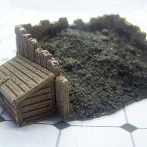 Pode incluir: Modelo em miniatura de um galpão e cerca de madeira desgastada, com um solo escuro e texturizado. O galpão tem um telhado inclinado e grão de madeira detalhado. A cerca é feita de tábuas verticais. O solo é um material escuro e granular.