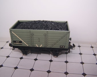 May include: A green toy train car with black wheels and a load of black coal. The car has the text "12T M608344 7-3" printed on the side.