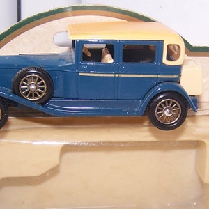 Puede incluir: Un modelo de coche de juguete vintage azul y crema. El coche tiene una parrilla negra y ruedas negras con detalles dorados.
