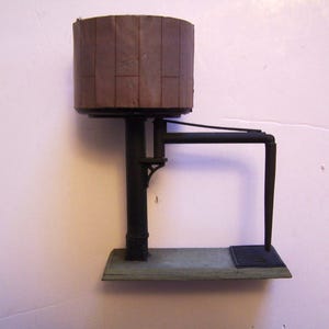 Pode incluir: Uma torre de água de trem modelo com um tanque de madeira marrom e suportes de metal preto. A torre tem um cano que se estende do tanque até o chão.