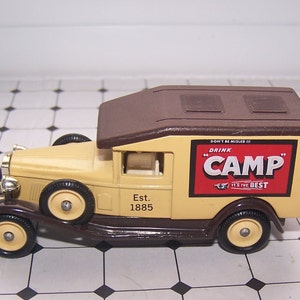 May include: A vintage toy delivery truck with a brown roof and a tan body. The truck has a "Drink Camp" sign on the side with the text "Don't be misled!! It's the best". The truck has an "Est. 1885" inscription on the side.