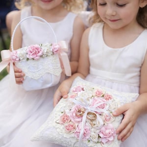 Pode incluir: Duas meninas em vestidos brancos seguram uma cesta de flores e uma almofada de anel. A cesta e a almofada são decoradas com flores rosa e brancas, renda e fitas. Anéis de casamento dourados estão na almofada.