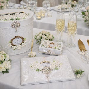 Op de afbeelding: Een verzameling bruiloftaccessoires, waaronder een witte kaartenbox, gastenboek en champagneglazen, allemaal versierd met kant, bloemen en sierlijke details. Gouden taartservers en een pen zijn ook aanwezig.