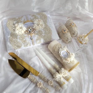 May include: An assortment of ivory and gold wedding accessories, including a guest book, champagne flutes, cake knife and server, candles, and a cake topper. The items are adorned with lace, pearls, and floral details, creating an elegant and romantic aesthetic.
