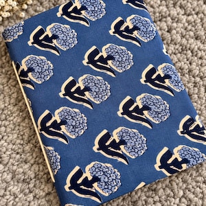 May include: A rectangular notebook with a blue fabric cover featuring a repeating pattern of light blue hydrangea flowers and dark navy blue stems. The notebook includes a matching blue tassel attached to the bottom edge of the spine.