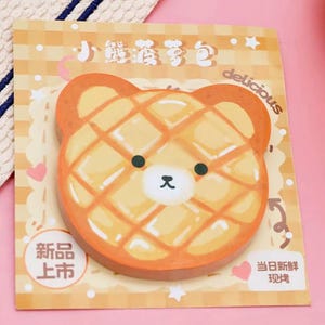 Könnte beinhalten: Ein bärenförmiger Haftnotizblock mit Waffelmuster in Orange- und Gelbtönen. Der Notizblock hat ein weißes Gesicht mit schwarzen Augen und einer kleinen schwarzen Nase. Die Wörter "delicious", "neues Produkt" und anderer Text sind sichtbar.