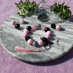 May include: A pink and black beaded bracelet with a pink tassel charm. The bracelet is made with various beads, including pink, black, and leopard print.