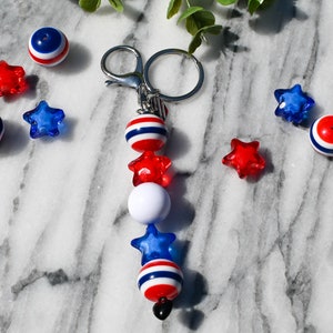 May include: A red, white, and blue beaded keychain with a silver key ring. The beads are in the shape of stars and circles. There are also loose red, white, and blue beads scattered around the keychain.