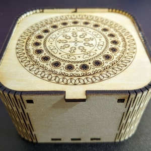 May include: A wooden box with a decorative mandala design on the lid. The mandala features a circular pattern with intricate details and a central motif of stylised symbols.