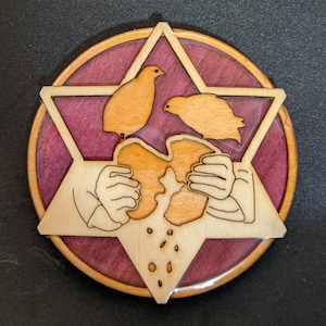 May include: A wooden coaster with a purple and brown background. A six-pointed star is cut out of the wood and features two birds and a hand holding a piece of bread.