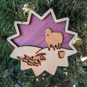 May include: A wooden Christmas ornament shaped like a star with a purple background. The ornament features a cutout of a sheep standing on a hill with a cup of coffee. A bee is flying towards the sheep.