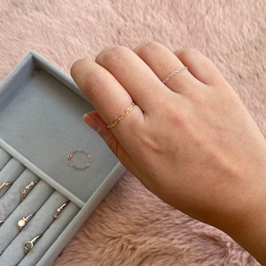 May include: Two gold chain rings on a finger. The rings are delicate and simple. The rings are in a ring box with other rings.