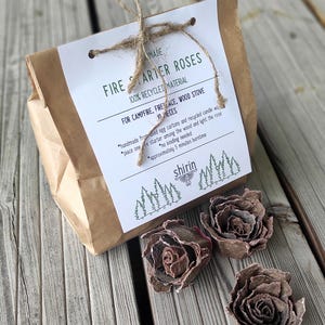 Könnte beinhalten: Eine braune Papiertüte, mit Schnur gebunden, mit der Aufschrift "FIRE STARTER ROSES" mit Text. Drei handgefertigte Feueranzünder in Rosenform liegen neben der Tüte. Die Feueranzünder bestehen aus recycelten Materialien.