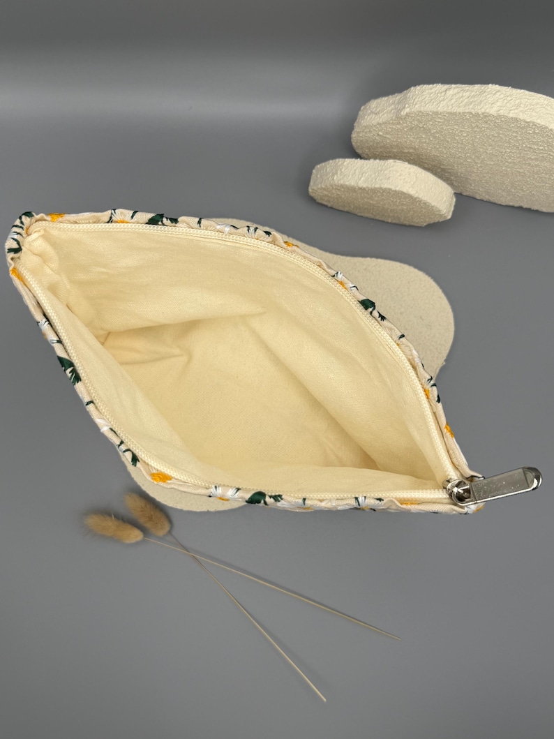 May include: A zippered pouch with a daisy floral pattern on a cream background. The interior is a light yellow color. The zipper is silver. The pouch is open, revealing the inside. Dried flowers are in the foreground.