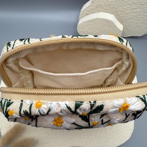 May include: A beige rectangular zippered pouch with an embroidered daisy pattern. The pouch features white daisies with yellow centers and green leaves. The interior is a light beige fabric with small pockets. The zipper is a gold color.
