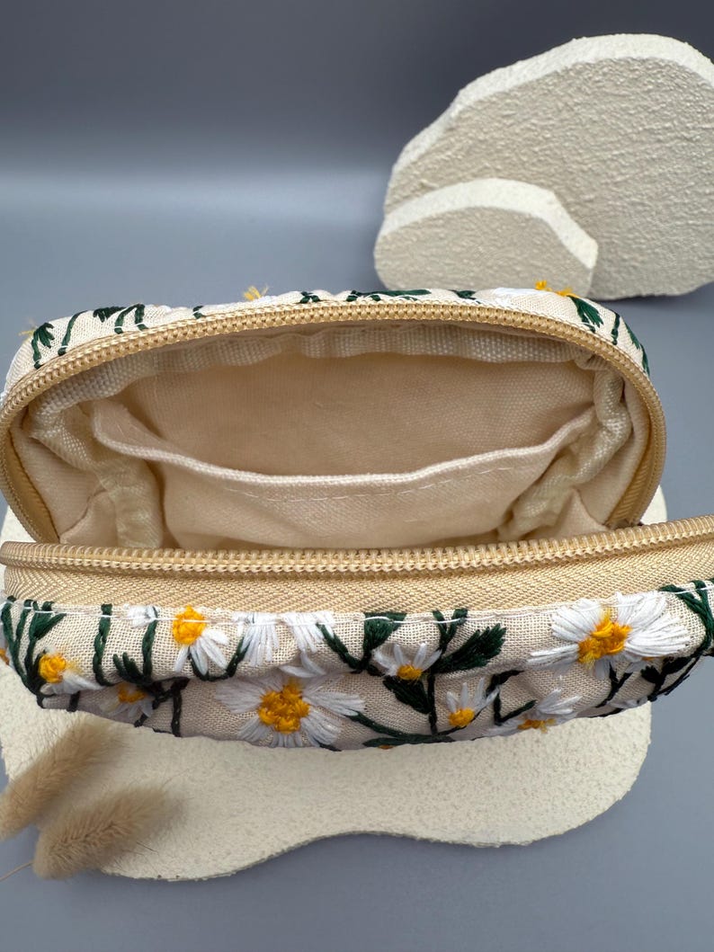 May include: A beige rectangular zippered pouch with an embroidered daisy pattern. The pouch features white daisies with yellow centers and green leaves. The interior is a light beige fabric with small pockets. The zipper is a gold color.