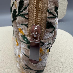 May include: Close-up of a beige fabric pouch with a floral daisy embroidery design. The pouch features a gold zipper and a silver zipper pull. The daisies are white with yellow centers and green leaves.