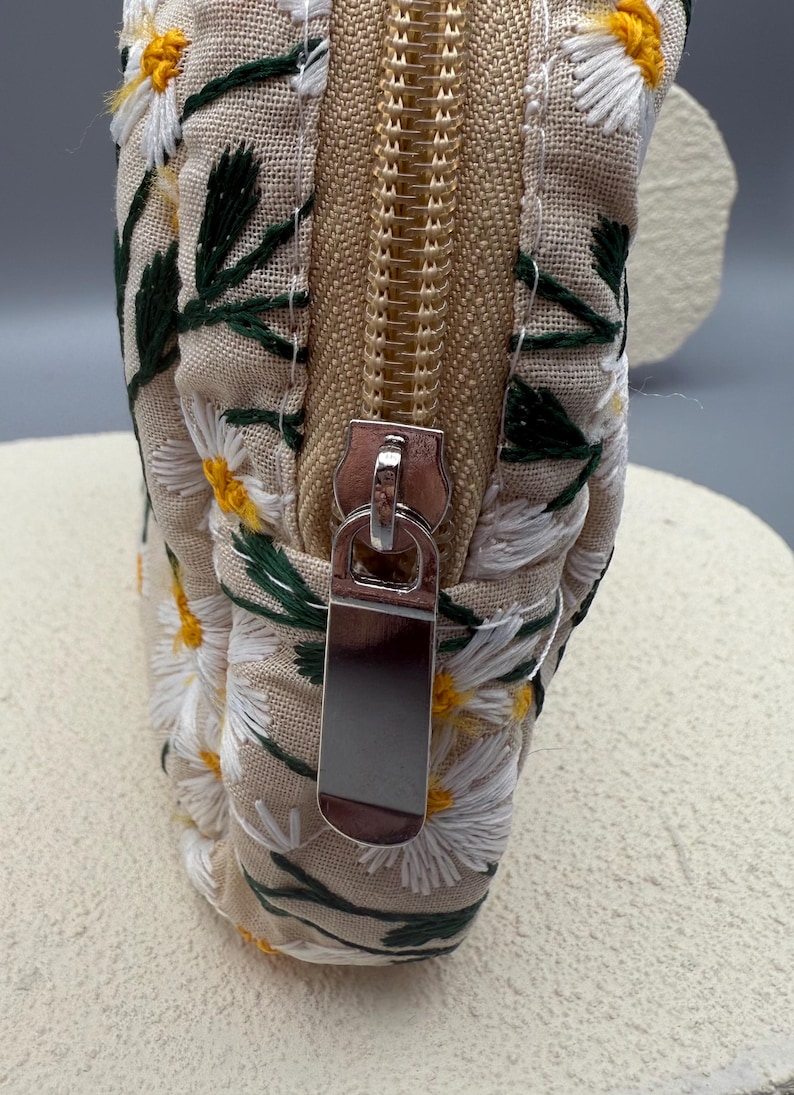 May include: Close-up of a beige fabric pouch with a floral daisy embroidery design. The pouch features a gold zipper and a silver zipper pull. The daisies are white with yellow centers and green leaves.