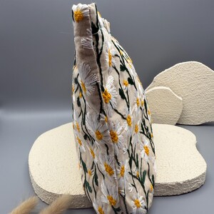 May include: A beige fabric pouch embroidered with a daisy pattern. The daisies have white petals and yellow centers, with green stems and leaves. The pouch is a soft, rectangular shape.