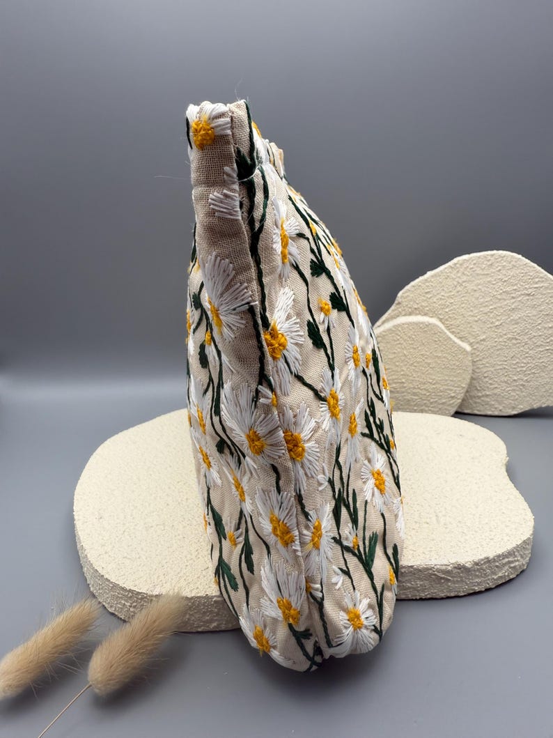 May include: A beige fabric pouch embroidered with a daisy pattern. The daisies have white petals and yellow centers, with green stems and leaves. The pouch is a soft, rectangular shape.