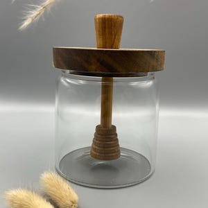 Könnte beinhalten: Ein klares Glas Honigglas mit einem Holzdeckel und einem Holzhonigrührer. Der Rührer hat ein Wabenmuster.