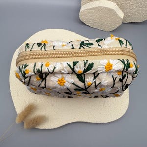 May include: A beige rectangular cosmetic bag with a daisy floral embroidery design. The bag features a gold zipper closure and is decorated with white daisies with yellow centers and green leaves.