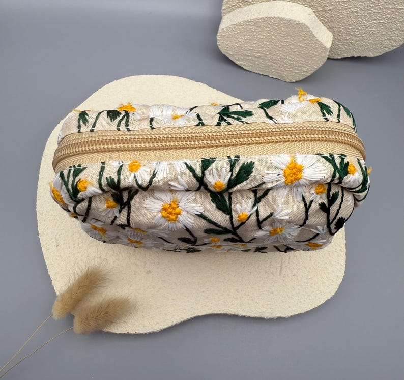 May include: A beige rectangular cosmetic bag with a daisy floral embroidery design. The bag features a gold zipper closure and is decorated with white daisies with yellow centers and green leaves.